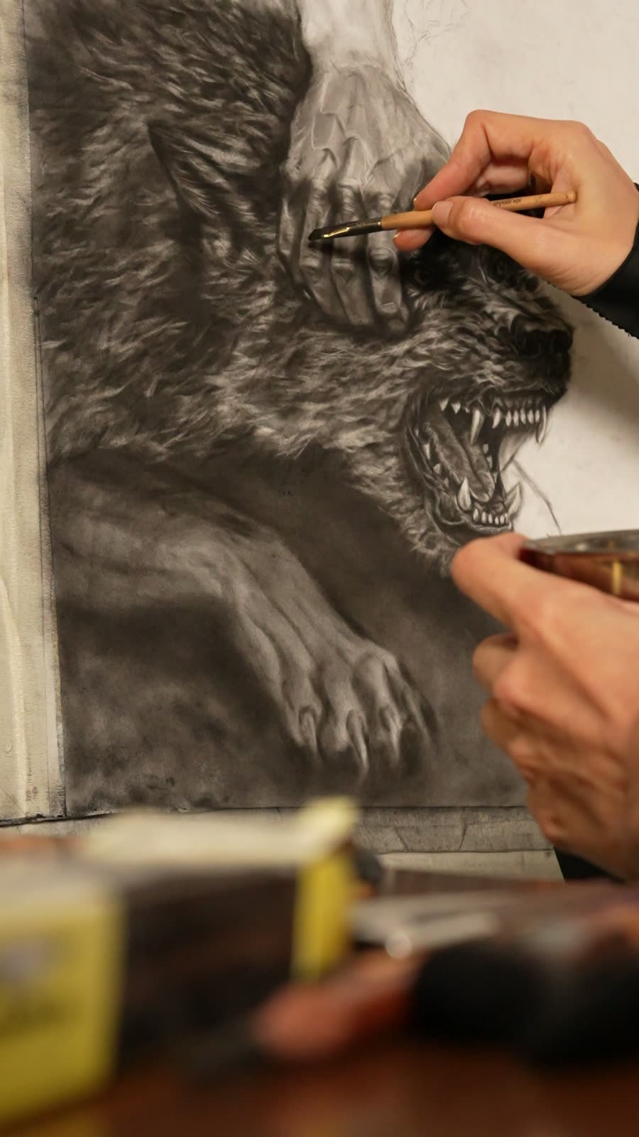 La obra en proceso: el lobo a carboncillo con las fauces abiertas, y la mano de Mariela trabajando sobre él con un difumino.